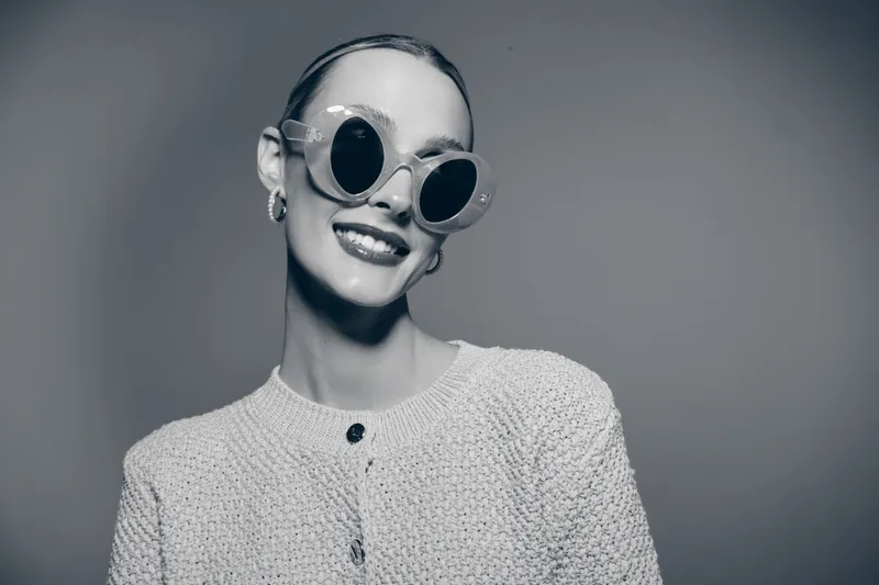 Portrait of a smiling young Caucasian woman with pale skin wearing round sunglasses and knitted sweater on grey background.