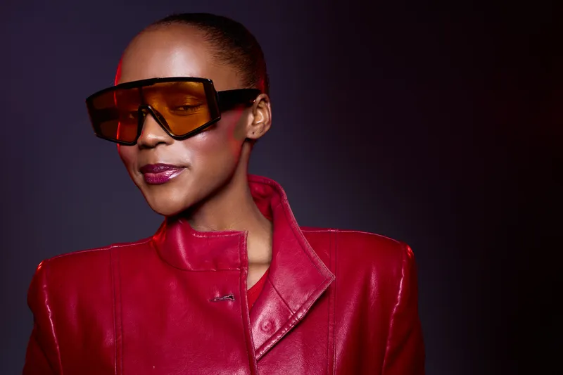 Confident African American woman with dark skin wears oversized orange sunglasses and a red leather jacket. Studio portrait with dramatic lighting, fashion concept and modern style.