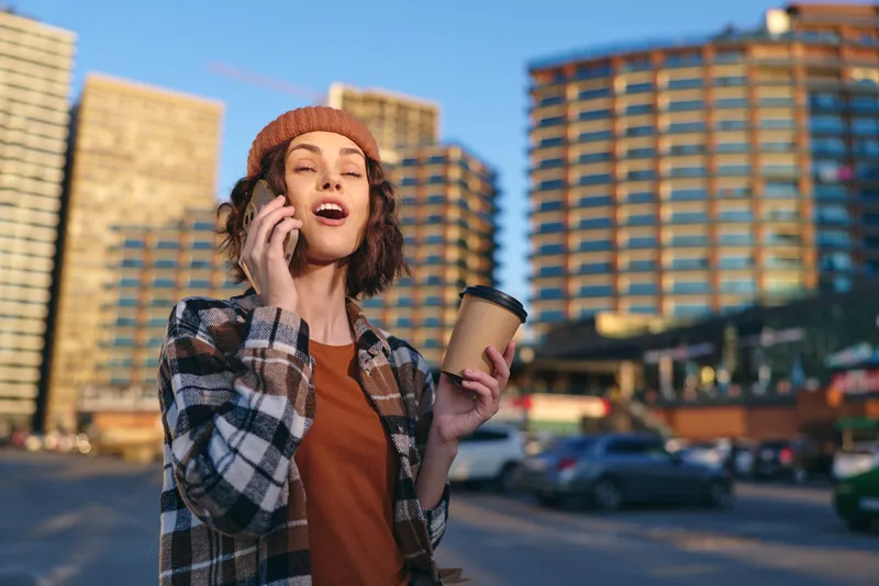woman phone coffee urban beanie smile in candid lifestyle scene of a young woman talking on phone with takeaway cup at golden hour glow, authenticity and emotional storytelling for mindful living