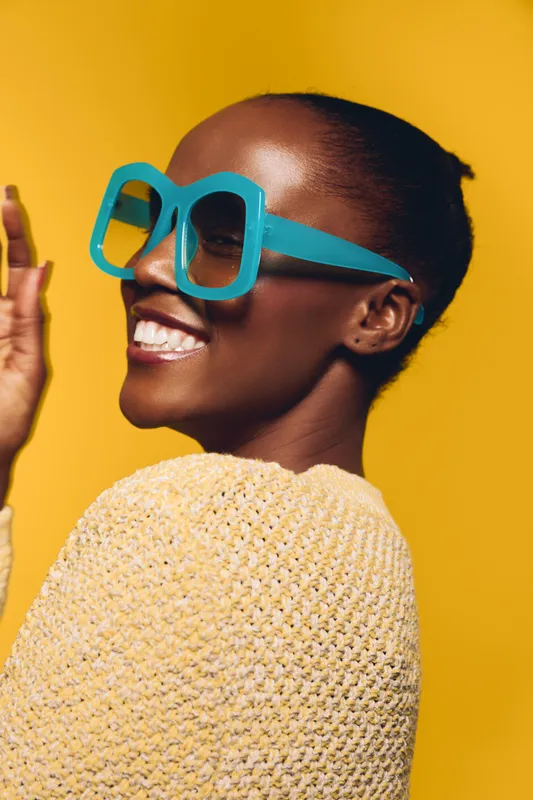 Happy black African woman with dark skin wearing large blue glasses and yellow sweater smiles joyfully against bright yellow background. Concept of fashion, style, and positive mood.