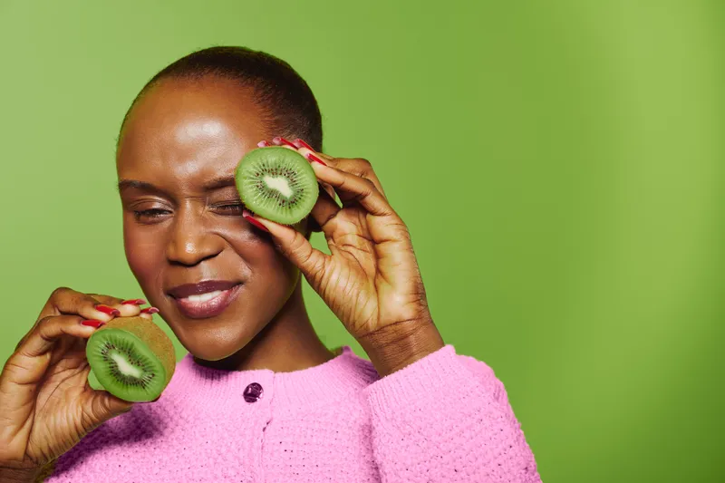 Dark skinned African woman in pink sweater holds two halves of fresh kiwi fruit near her face with one eye closed. Bright green background and natural skincare concept with healthy lifestyle mood.