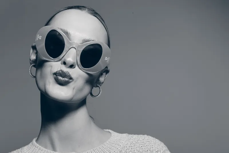Stylish bald white woman with pale skin wearing large round sunglasses and hoop earrings poses with puckered lips in studio. Concept of fashion, bold look, and confidence.