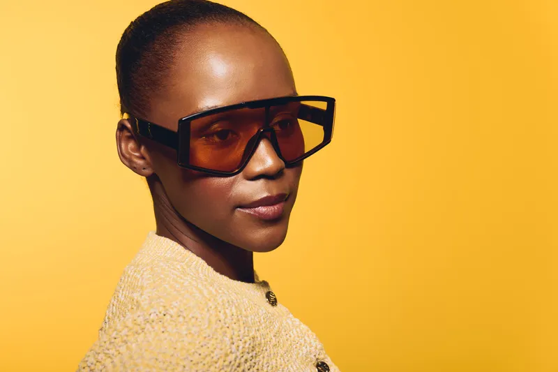 Confident African woman with dark skin wearing oversized sunglasses and a yellow knitted sweater poses against a bright yellow background. Concept of fashion, style, and beauty.