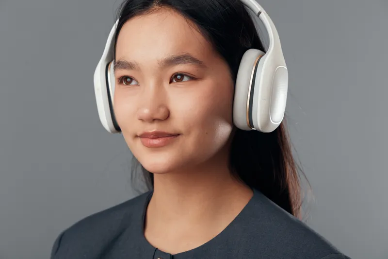 Young woman with headphones wearing dark blouse listens to music with eyes looking away on grey background. Attractive female portrait with long hair and calm expression closeup studio shot.