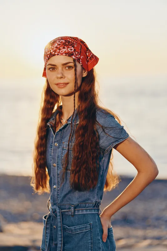 lifestyle woman in modern boho-western denim outfit with long hair and red bandana, standing on beach during golden hour, film color style lighting