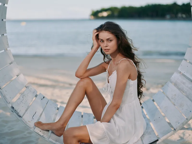A young woman sits on a wooden swing at the beach, wearing a light white dress. Soft waves lap the shore as a calm seascape frames a gentle, intimate portrait.