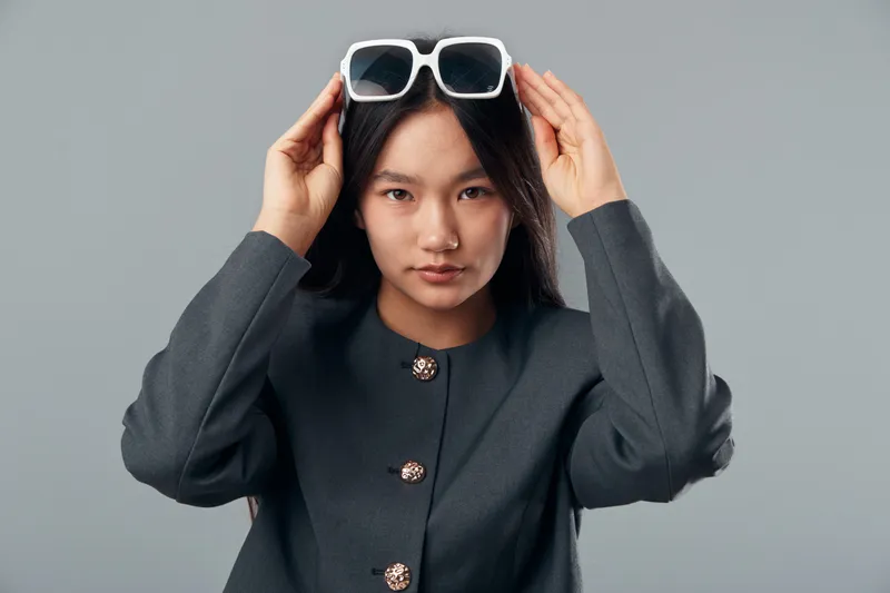 Woman posing with sunglasses on head wearing dark grey coat with button details against neutral background for fashion style and confident portrait in studio.