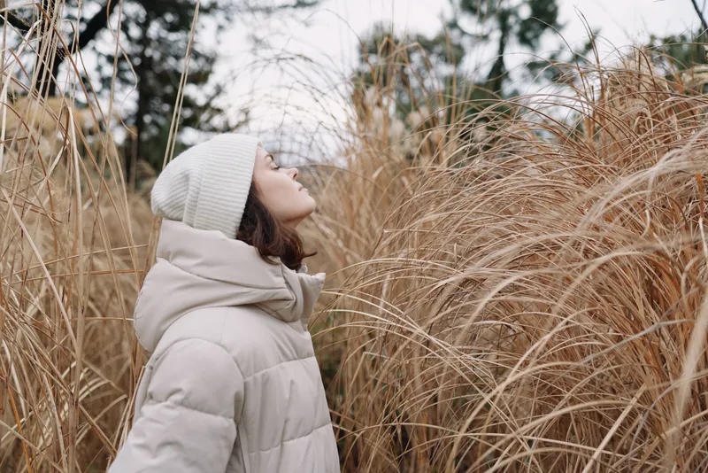 Woman in a beige winter coat and scarf amid tall dried grasses, gazing upward in a calm outdoor nature scene with soft light and seasonal atmosphere.