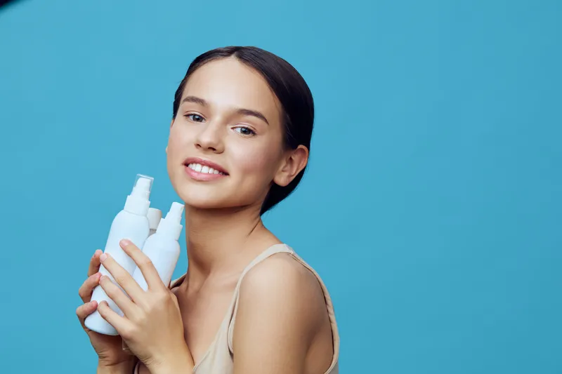 Young girl holding skincare bottles, smiling happily against a vibrant blue background, showcasing youthful beauty and healthy skincare routine