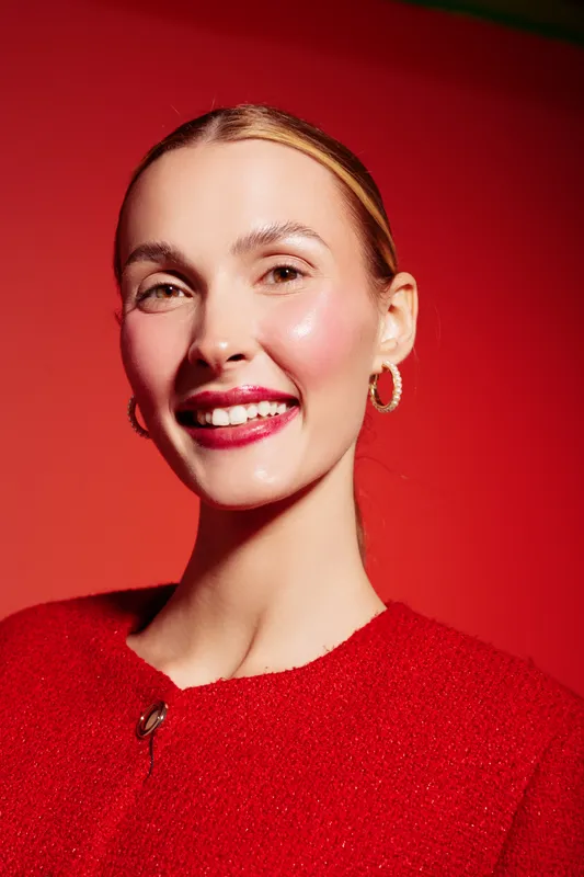 Smiling woman with red lipstick and hoop earrings poses against a red background wearing a red textured top. Concept of beauty, fashion, confidence, and vibrant style portrait.