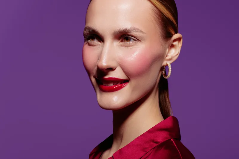 Close-up portrait of a smiling woman with red lipstick and hoop earrings against a purple background. Concept of beauty, fashion, makeup, and confident female model in studio lighting.