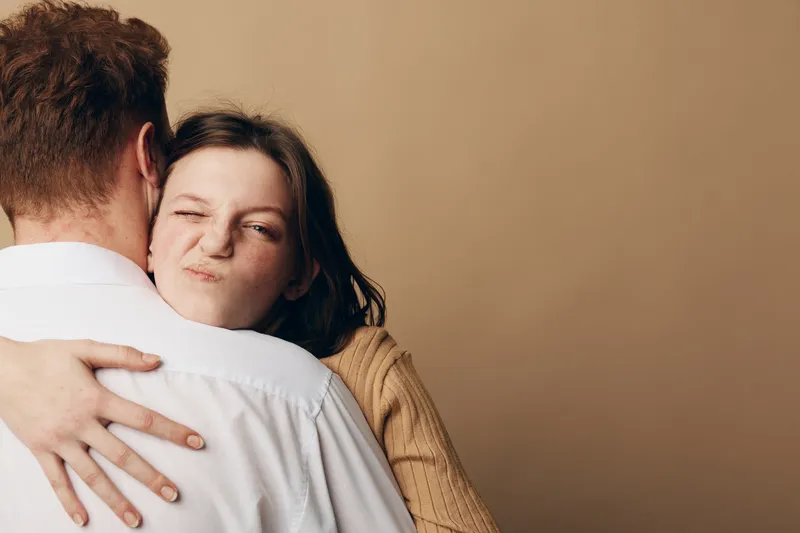 Young woman in brown sweater hugs man in white shirt, making a playful face. Intimate moment with warm beige background, concept of affection, relationship, and casual lifestyle.