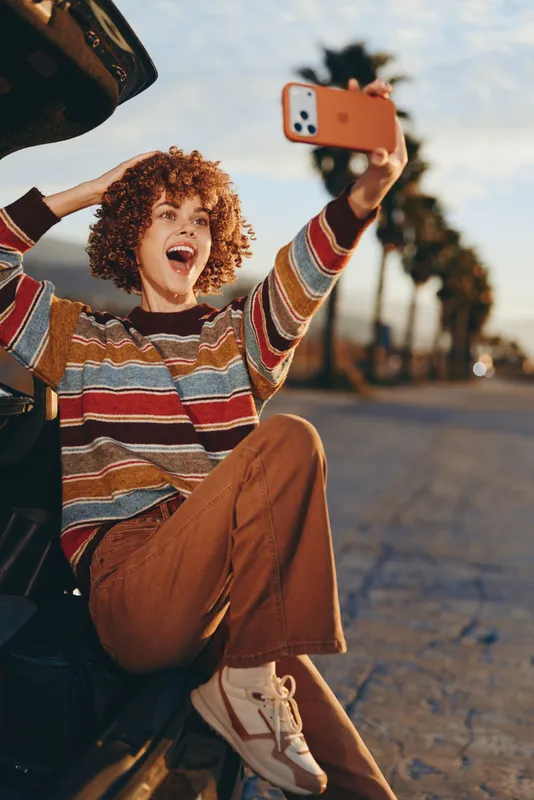 Woman selfie with smartphone outdoors on street, curly hair, casual fashion striped sweater and brown pants, laughter joy in lifestyle moment on sunny road, travel vibe