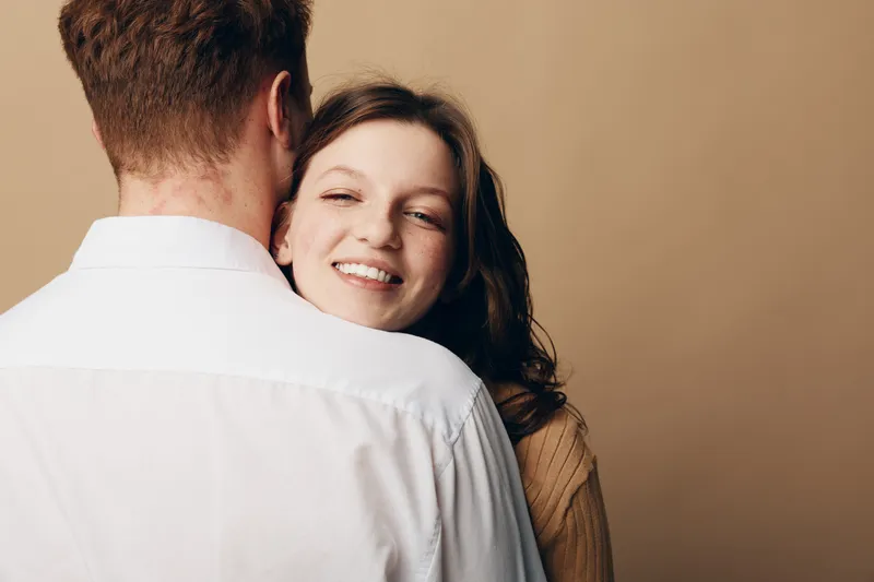 Smiling woman with long hair embraces man wearing white shirt on beige background. Concept of love, relationship, happiness, and intimacy between couple in casual clothing.
