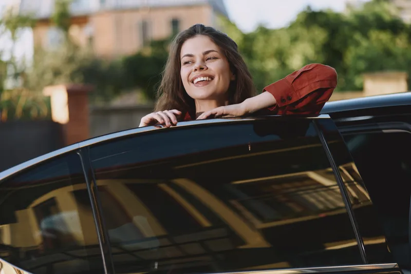 Smiling woman leaning on a car window in sunlight, showcasing a casual lifestyle moment outdoors, with cheerful, relaxed, carefree mood and summer energy