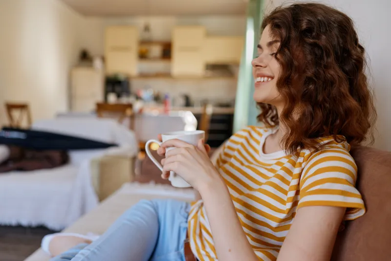 Young woman with wavy hair relaxes on a cozy couch indoors, smiling while holding a mug, enjoying a casual home moment under soft natural light