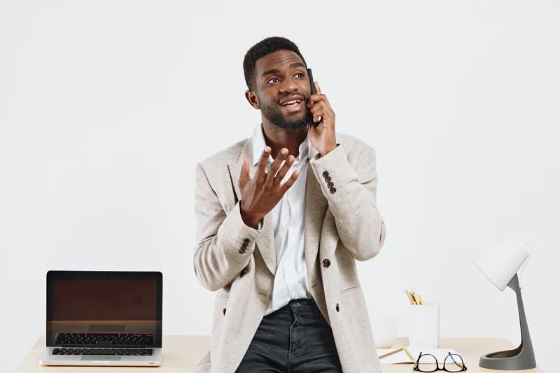 Smiling young African-American man wearing beige cardigan talking on mobile phone in office with open laptop and accessories on desk, looking happy and engaged. People lifestyle concept.