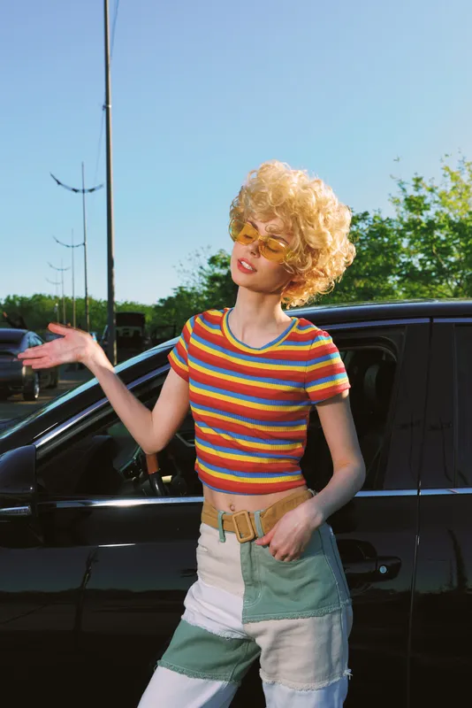 Cheerful woman outdoors, retro fashion with a striped crop top and patchwork shorts, curly hair, sunlit moment, casual pose beside a car, vibrant colors, natural light, carefree vibe