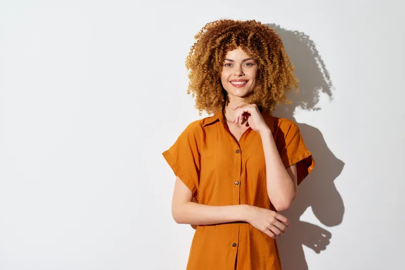 Улыбающаяся молодая женщина с объёмными вьющимися волосами, одетая в коричневую рубашку, позирует на фоне однотонного белого фона