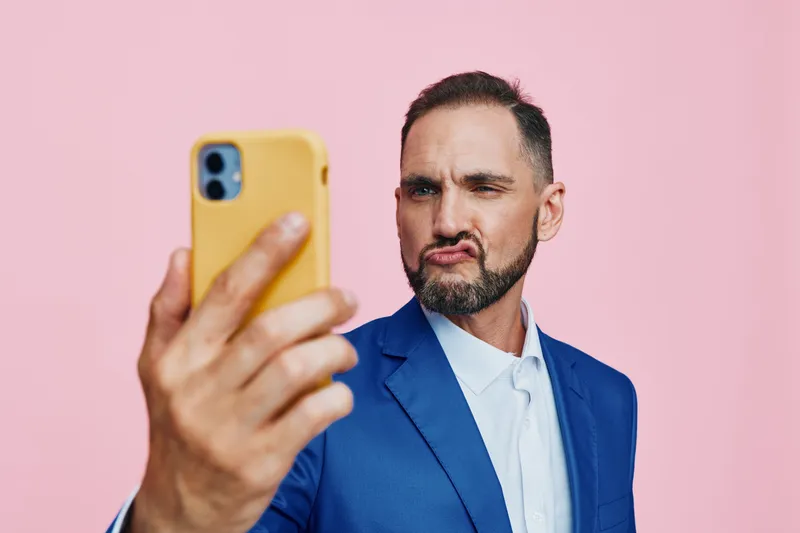 A professional man in a blue suit takes a confident selfie against a solid pink background, showcasing modern business style and a determined expression.
