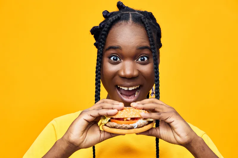 Happy young Black woman excitedly holding a delicious hamburger against a vibrant yellow background, showcasing joy and a sense of indulgence in fast food