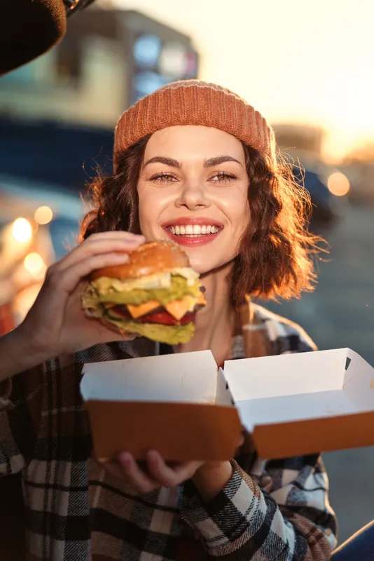Woman with beanie and plaid jacket smiling while holding burger outdoors at golden hour, candid lifestyle portrait evoking authenticity, mindful living and emotional storytelling in urban sunset.