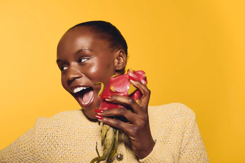 Happy black woman with dark skin laughs holding a dragon fruit near her face on yellow background. Concept of healthy lifestyle, exotic fruit, joy, natural beauty, and vibrant mood.