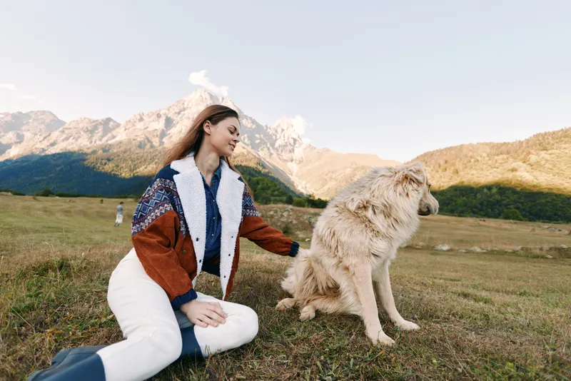 Woman petting a dog in a mountain meadow, outdoor portrait of pet and owner sitting on grass, smiling and enjoying nature in a cozy sweater, relaxed friendship moment.