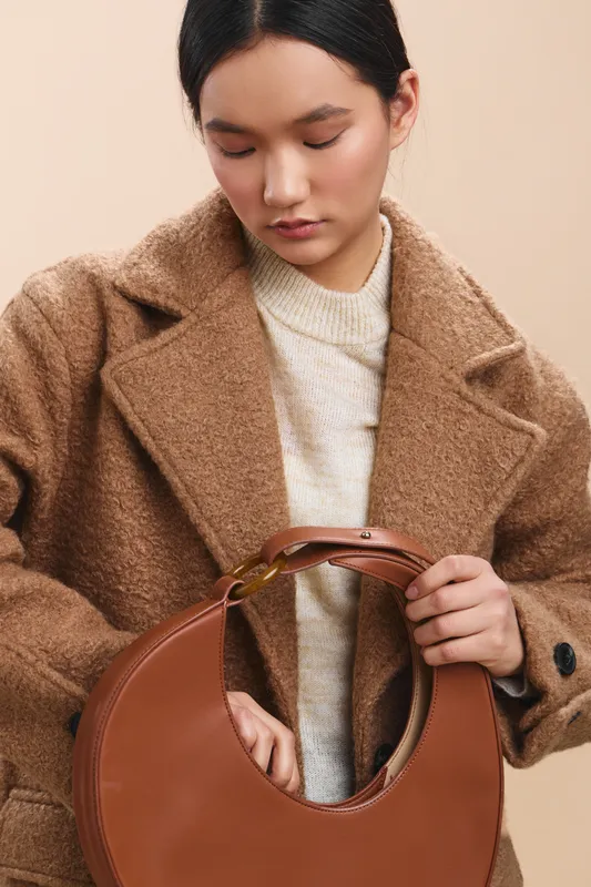 Asian woman wearing a tan coat examines a round brown handbag in a minimalist setting, exuding calm confidence and refined fashion sense, with soft lighting and a neutral backdrop that highlights