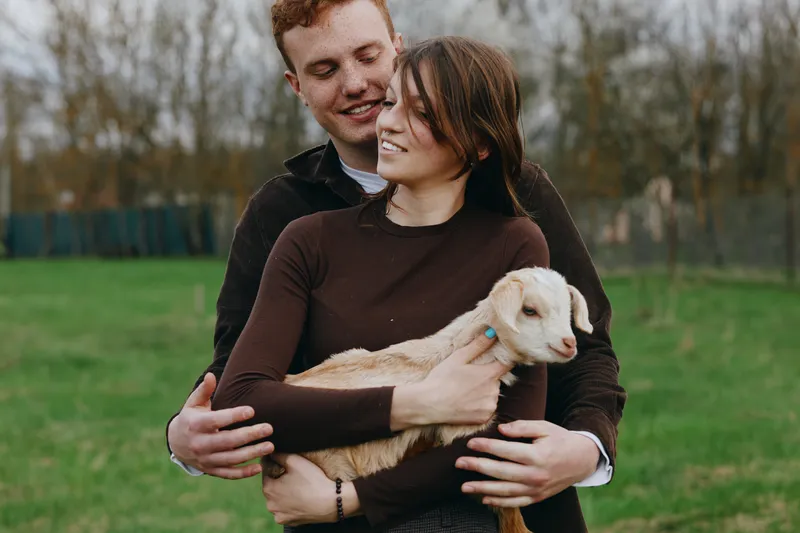 A couple in sweaters cradle a young goat in an open field, capturing a warm family moment. Natural light highlights bonding, calm countryside, and a rural lifestyle with a playful animal companion.