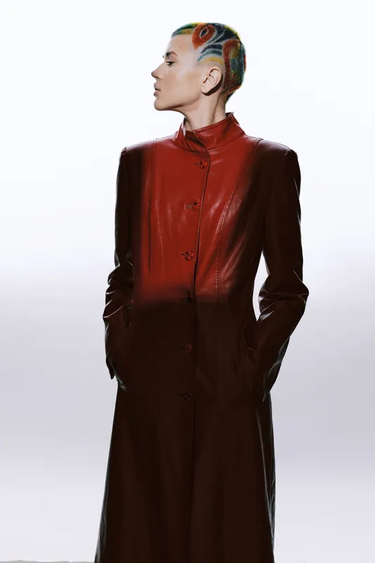 Woman with colorful shaved hair wearing a long red leather coat stands confidently against a white background, showcasing modern fashion and bold style.