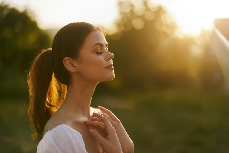 Serene young woman enjoying sunset glow in nature, wearing white dress, conveying peace and tranquility amidst greenery, perfect for wellness concepts