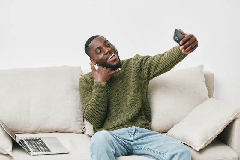 Young Black man smiling and taking a selfie on a couch with a laptop, wearing a green sweater, conveying joy and comfort at home