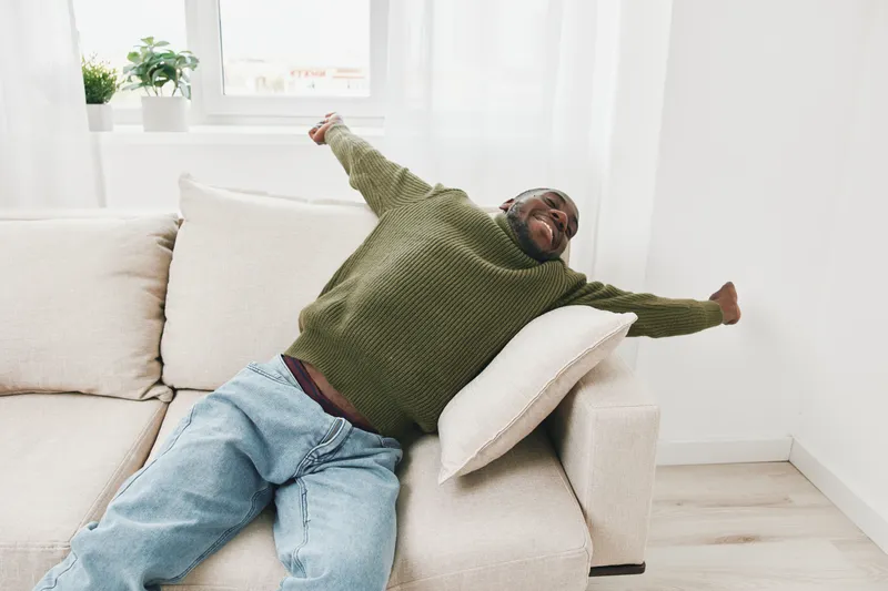 Happy black man relaxing on a couch in a cozy living room, wearing a green sweater and jeans, with an expression of joy and comfort