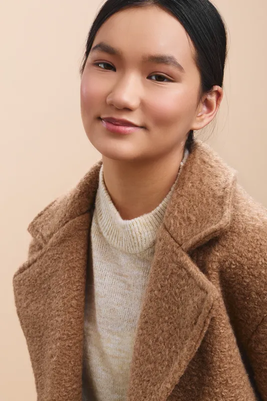 cheerful asian woman poses in a warm coat and knit sweater against a neutral backdrop, with soft lighting that highlights her calm smile and confident expression.