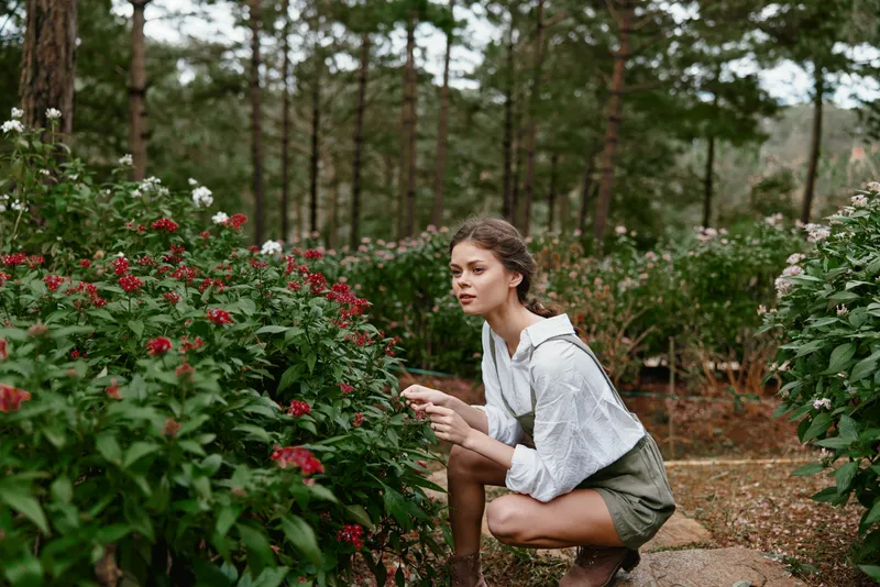Young woman in a light outfit exploring flowers in a lush garden, expressing curiosity and joy amidst vibrant greenery and colorful blooms