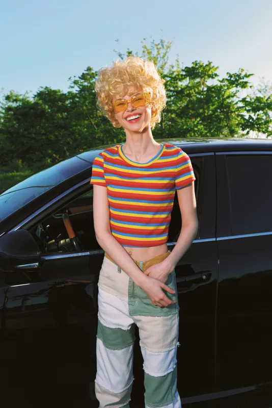 Young woman with curly blond hair in a striped crop top leans beside a black car in a sunny outdoor setting, capturing a playful retro film color mood and a bright cheerful vibe