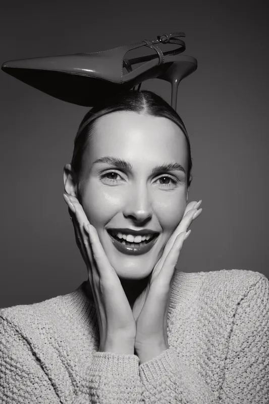 Smiling Caucasian woman with light skin wearing knitted sweater balances high heel shoe on head. Studio portrait with creative concept and playful mood.