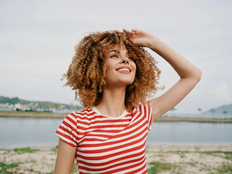 Smiling woman with curly hair wearing a red and white striped tee stands by a tranquil lakeside, sunlight warming her face as a gentle breeze lifts her smile.