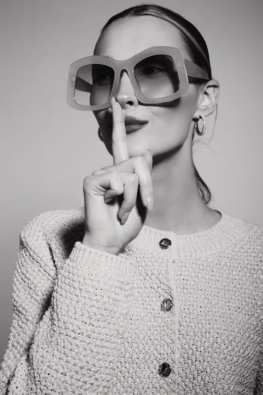 Elegant white woman with pale skin wears large sunglasses and textured jacket, finger on lips in gesture for silence or secret.