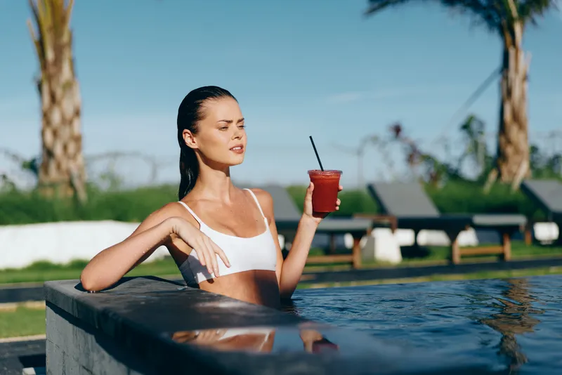 Cheerful woman by a pool, wearing a white top, sipping a red drink, sunlit water reflections, relaxed pose, tropical mood, lush greenery background