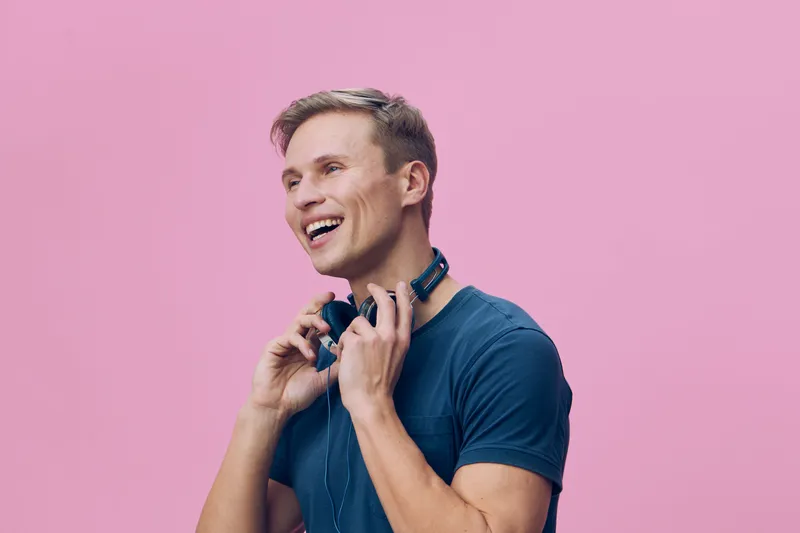 Smiling man wearing casual blue t-shirt holding headphones around neck look happily at camera studio portrait isolated on pink background cheerful joyful expression