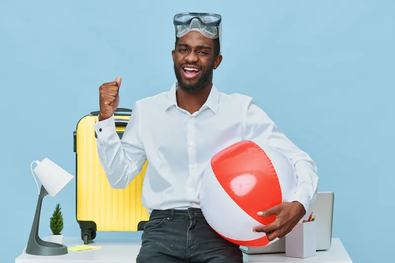 Happy young black man in casual shirt holding beach ball and raising fist in victory gesture, standing in travel accessories on blue studio background, summer vacation concept