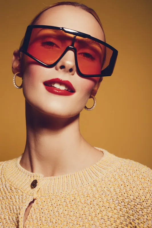 Stylish woman with oversized orange sunglasses and red lipstick poses confidently against a warm yellow background. Fashion concept with bold accessories and makeup.