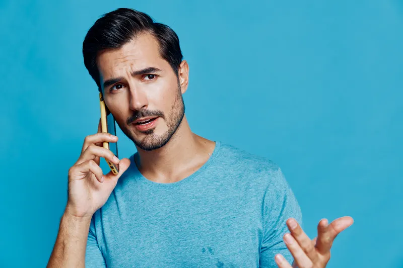 Concerned young man talking on mobile phone, wearing blue t-shirt, looking thoughtfully at camera against vibrant blue background. Communication lifestyle concept.