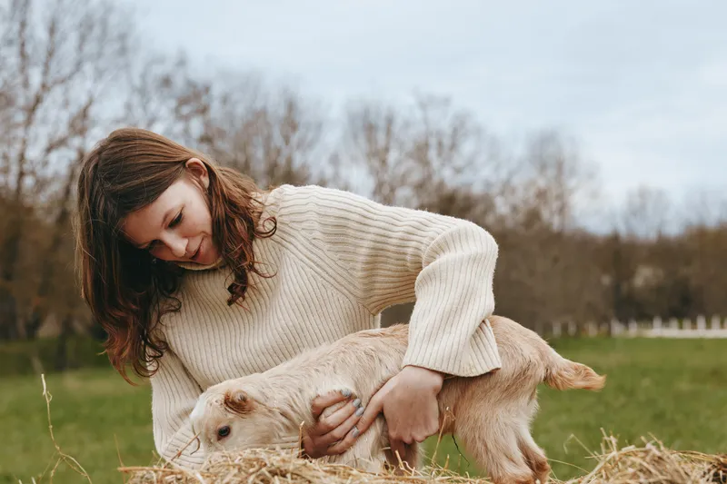 Cheerful woman outdoors, stroking a goat kid in a grassy field, cozy sweater, gentle breeze, natural colors, relaxed countryside moment, warm lighting, peaceful mood outdoor lifestyle