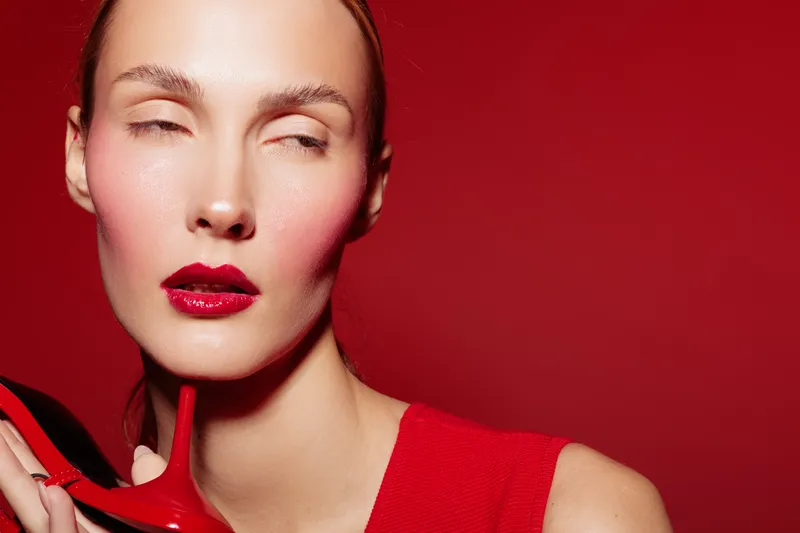 Close-up of a woman with red lipstick and blush holding a red high heel shoe near her face on a red background. Concept of fashion, beauty, and bold makeup style.