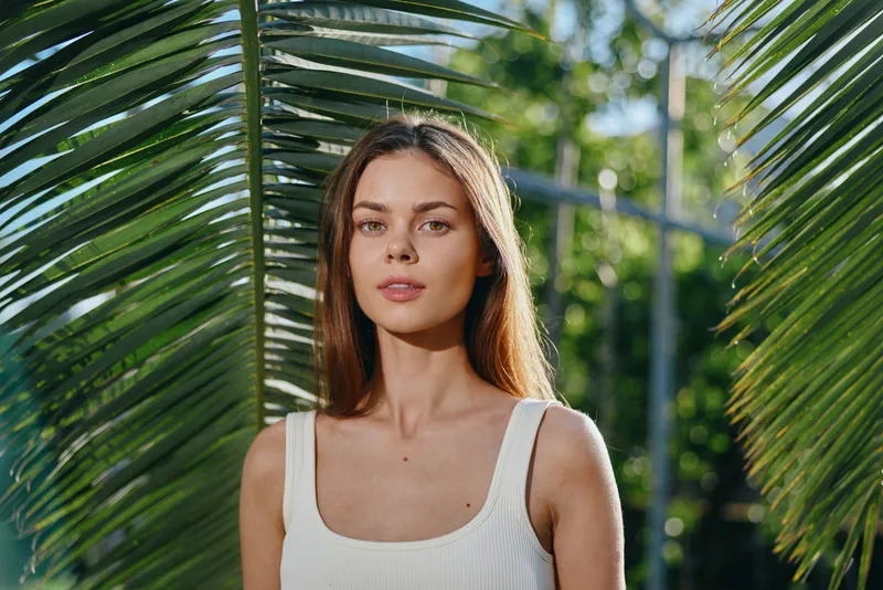 Young woman with long hair and natural makeup posing outdoors with green palm leaves and sunlight in summer, wearing a white tank top standing in tropical environment.