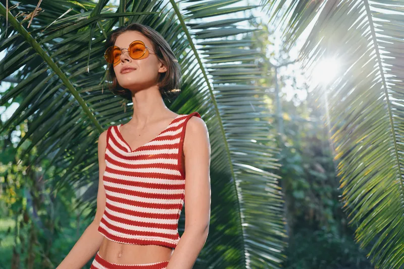 A stylish young woman stands outdoors among large tropical leaves, wearing a red and white striped crop top and sunglasses, exuding carefree summer vibe and sunny confidence.