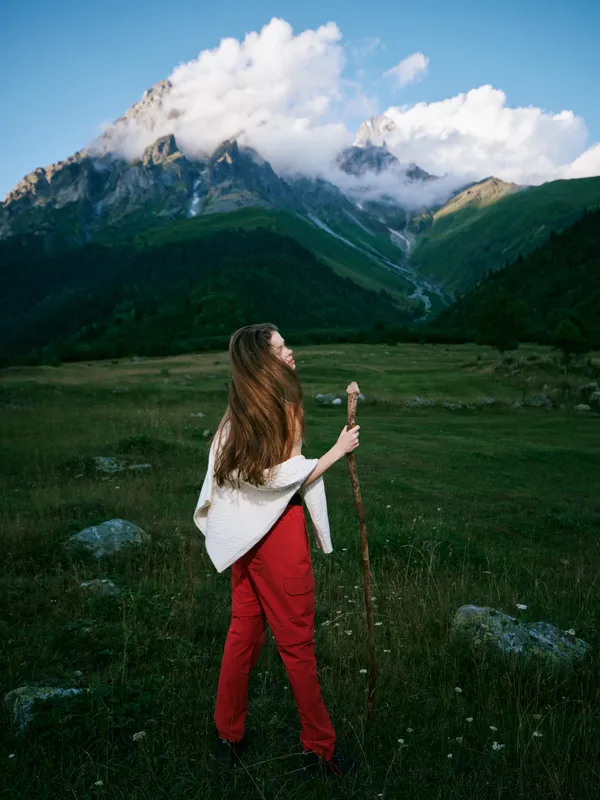 Woman in red pants holds a walking staff in a meadow, gazing toward towering mountains under a cloudy sky, conveying adventure, solitude, and outdoor exploration in pristine terrain.